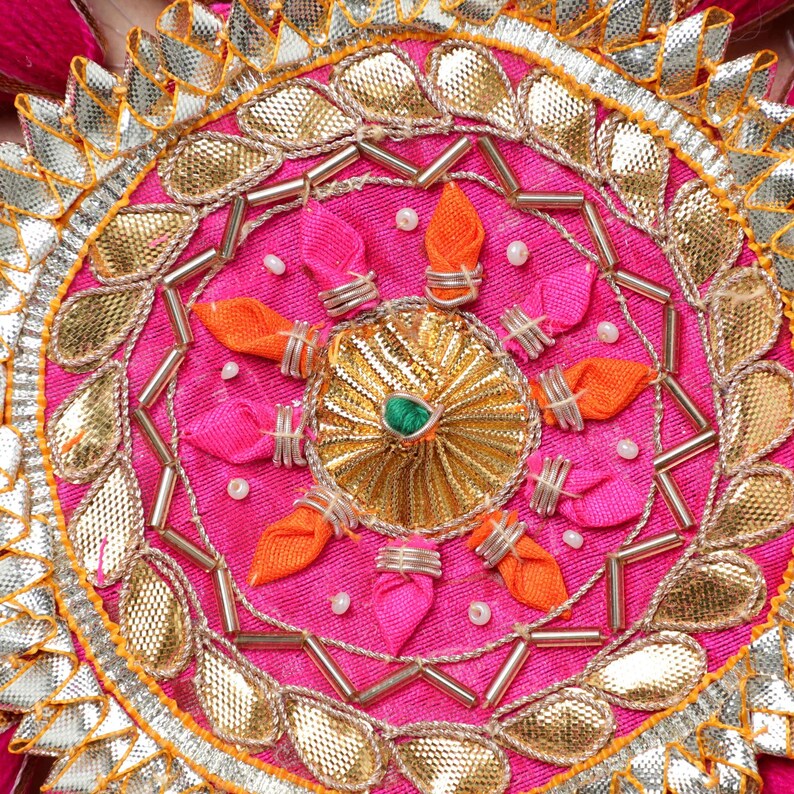 May include: A pink fabric circle with a gold and silver design. The center of the circle has a gold and green flower design. The circle is decorated with small pink and orange fabric flowers.
