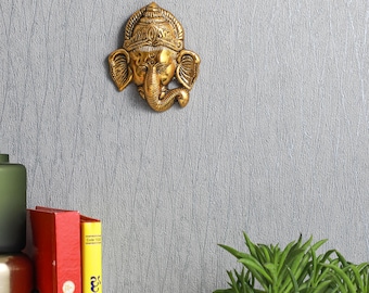 May include: A gold-colored metal wall hanging of the Hindu deity Ganesha. The sculpture is detailed with intricate carvings and features a traditional headdress.