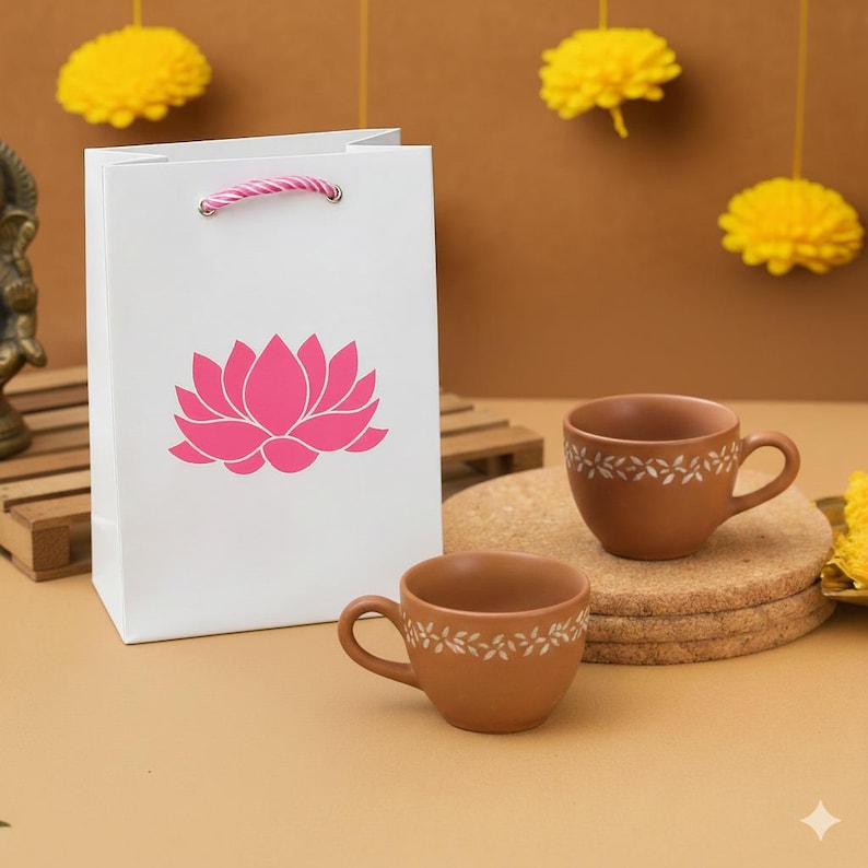 Puede incluir: Una bolsa de regalo blanca con un dise&ntilde;o de flor de loto rosa y un asa de cuerda rosa y blanca. Dos tazas de t&eacute; de cer&aacute;mica marr&oacute;n con dise&ntilde;os florales blancos est&aacute;n sobre un posavasos de corcho. Decoraciones de pomp&oacute;n amarillas cuelgan en el fondo.