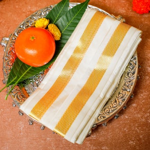 Op de afbeelding: Een witte zijden stof met gouden strepen, gevouwen en geplaatst op een zilveren dienblad met een mango en gele bloemen.