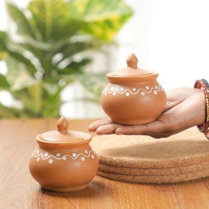 May include: Two small, round, terracotta-coloured pots with lids. Each pot has a white, wavy, dotted design around the base. One pot is held in a hand, while the other sits on a wooden surface. The background features a green plant.