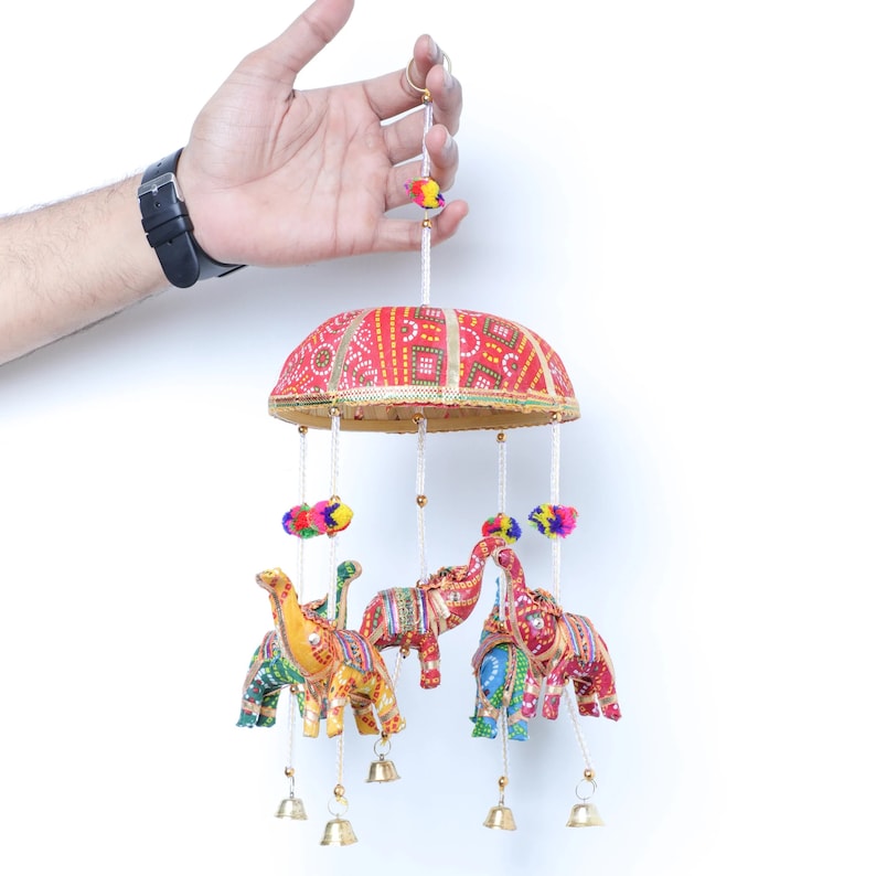 May include: A colorful hanging mobile with a red, yellow, and blue patterned fabric canopy and four hanging elephants. The elephants are decorated with colorful patterns and have bells hanging from their feet.