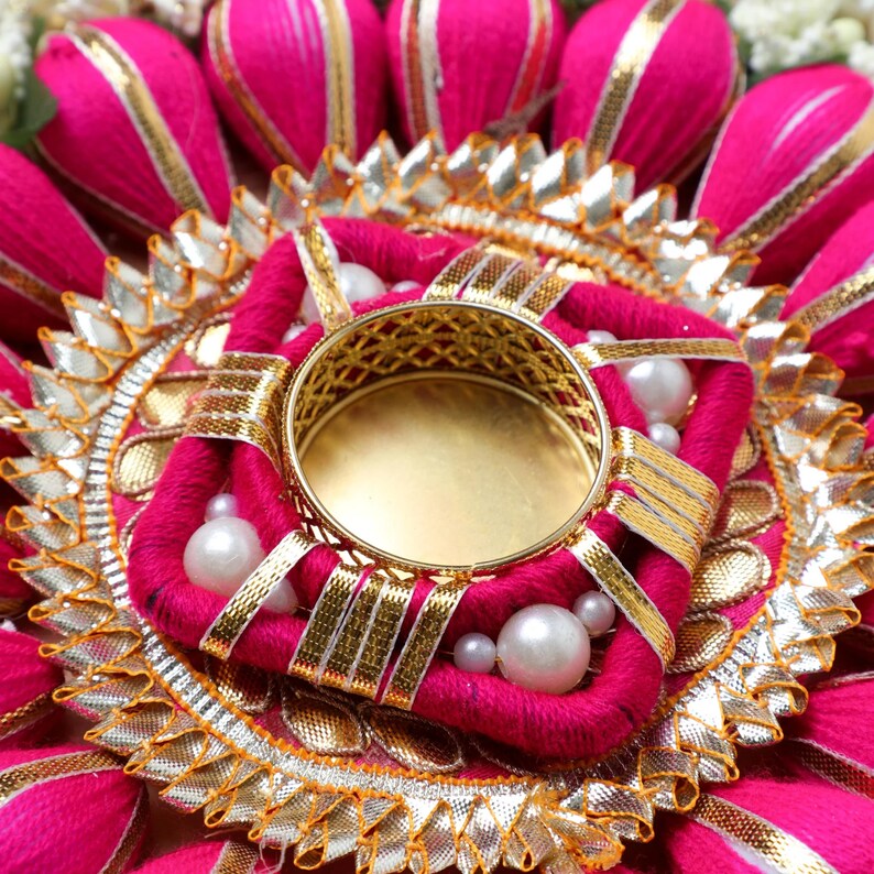 May include: A pink and gold decorative diya with a round metal holder in the center. The diya is decorated with white pearls and gold ribbon.