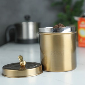 May include: A gold-colored tea canister with a silver rim, filled with loose tea. The lid is off, revealing the tea inside. The lid has a small bird-shaped handle. The word "chai" is etched on the side of the canister.