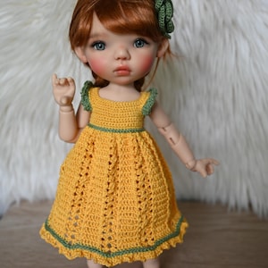 May include: A ball-jointed doll with auburn hair and a green hair accessory, wearing a yellow crocheted dress with green trim at the waist, hem, and sleeves, and matching green crocheted shoes. The doll has articulated limbs and painted facial features.