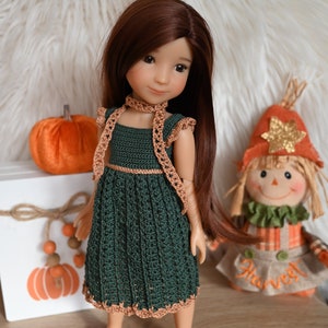 May include: A doll wearing a green crocheted dress with brown trim and a brown crocheted scarf. The doll has long brown hair and brown eyes. The doll is standing on a wooden surface with a small pumpkin and a scarecrow in the background.