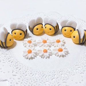May include: A set of six yellow and black fondant bumblebees with white wings, and six white fondant daisies with yellow centers. The bumblebees and daisies are arranged on a white doily.