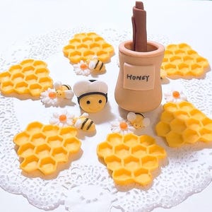 May include: A honey themed cake topper with a brown honey jar, a wooden stick, and yellow honeycomb decorations. The honey jar has the word "Honey" written on it. There are also small yellow bees and white flowers.