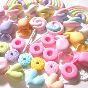 May include: A collection of pastel-colored candies, including donuts, lollipops, and heart-shaped sweets. The image features a variety of shapes and colors, such as pink, blue, yellow, and purple, arranged on a white surface. The candies are likely handmade.