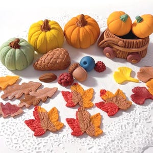 May include: An assortment of fall-themed decorations, including pumpkins, leaves, acorns, and a basket with pumpkins. The pumpkins are in shades of yellow and orange, and the leaves are in various autumn colors. The decorations are arranged on a white, lacy surface.