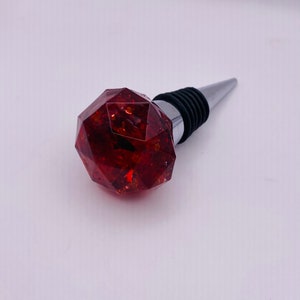 May include: A decorative wine bottle stopper with a faceted, reddish-brown gem-like top. The stopper has a silver-colored metal base and a black rubber seal. The gem-like top has a geometric shape and appears to have inclusions.