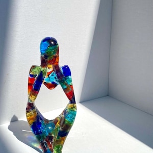 May include: A colorful glass sculpture of a person sitting with their arms crossed in front of them. The sculpture is made of many small pieces of glass in various colors, including blue, green, red, yellow, and orange.
