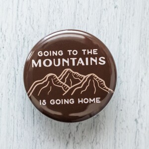 May include: A brown button with white text that reads "Going to the Mountains is going home". The button features a line drawing of three mountains in a light brown color.
