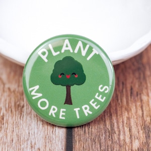 May include: A round, green button with the words "PLANT MORE TREES" in white lettering. The button features a cartoon tree with a smiling face. The button is on a wooden surface.