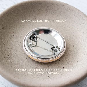 Pu&ograve; includere: Il retro di un bottone da spilla da 3,17 cm con una spilla argentata e un retro bianco. Il testo "EXAMPLE 1.25 INCH PINBACK" e "DOTTIEANDCARD.COM" sono stampati in oro sul retro. Il testo "ACTUAL COLOR VARIES DEPENDING ON BUTTON DESIGN" &egrave; stampato in oro su una superficie marrone chiaro.