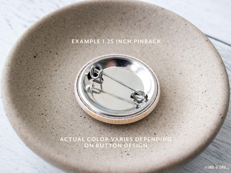 Pu&ograve; includere: Il retro di un bottone da spilla da 3,17 cm con una spilla argentata e un retro bianco. Il testo "EXAMPLE 1.25 INCH PINBACK" e "DOTTIEANDCARD.COM" sono stampati in oro sul retro. Il testo "ACTUAL COLOR VARIES DEPENDING ON BUTTON DESIGN" &egrave; stampato in oro su una superficie marrone chiaro.