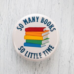 May include: A white button with blue text that reads "So many books, so little time". A stack of rainbow colored books is in the center of the button.