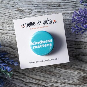 Pu&ograve; includere: Un bottone da spilla blu con il testo "kindness matters" in bianco. Il bottone &egrave; su una carta bianca con il testo "Dottie & Caro" e "Pinback Button" in nero.