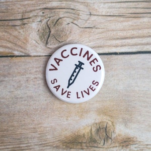 May include: White button with the text "Vaccines Save Lives" in maroon lettering and a black silhouette of a syringe in the center.