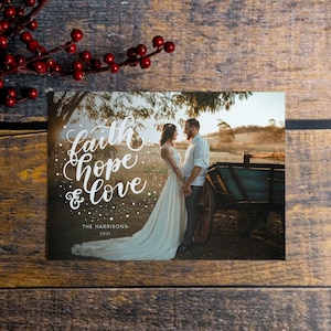 May include: A Christmas card with a photo of a couple standing in a field with a wagon in the background. The card has the words "faith hope & love" written in white script on a snowy background. The text "The Harrisons 2021" is printed below the words.