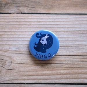 May include: A blue button with a stylized illustration of a woman's face with a flower in her hair. The text "VIRGO" is printed below the illustration.