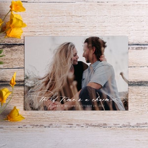 May include: A wedding announcement card with a photo of a couple looking at each other. The text on the card reads "Third time's a charm" with the couple's names and wedding date below.