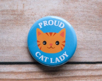 May include: A blue button with a white border and the text "Proud Cat Lady" in white. A cartoon orange tabby cat with black stripes is in the center of the button.