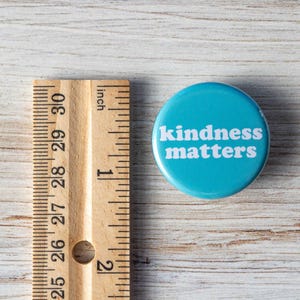 Pu&ograve; includere: Un bottone blu con il testo "kindness matters" in lettere bianche. Il bottone ha un diametro di circa 2,5 cm.