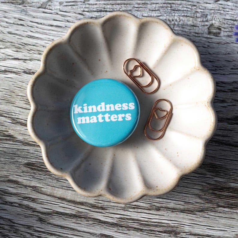 Pu&ograve; includere: Un bottone blu con il testo "kindness matters" in lettere bianche, posto su un piatto di ceramica bianca con due graffette a forma di cuore dorate.