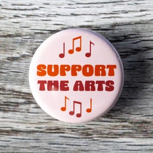 May include: A pink button with the text "SUPPORT THE ARTS" in orange and red letters. There are musical notes in orange and red on the top and bottom of the button.