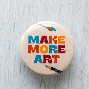May include: A round button with the text "MAKE MORE ART" in colorful letters. The button has a white background and features two paintbrushes, one with a blue tip and the other with a black tip.