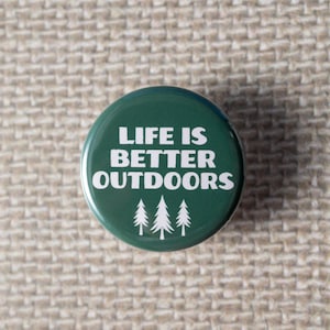 May include: A round, forest green button with the white text "LIFE IS BETTER OUTDOORS" and three white pine trees. The button is set against a textured, beige background.