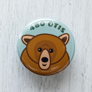 May include: A blue and green button with the number 480 and the word "OTIS" printed on it. A cartoon brown bear face is in the center of the button.