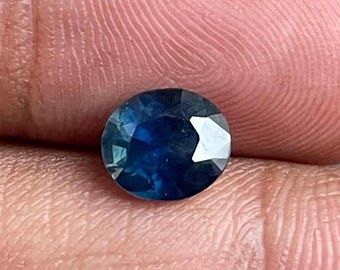 May include: A faceted, round, deep blue gemstone. The gem is held between fingers, showcasing its brilliant cut and color. The stone's facets reflect light, creating a sparkling effect. This gemstone is suitable for jewelry making.