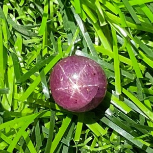 May include: A round, polished, purplish-red gemstone with a six-rayed star effect, resting on a bed of bright green artificial grass. The gemstone's surface reflects light, creating a star-like pattern.