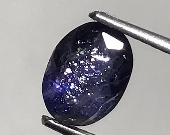 May include: A dark blue oval-shaped gemstone with a sparkly, iridescent finish. The gemstone is held by a pair of tweezers.