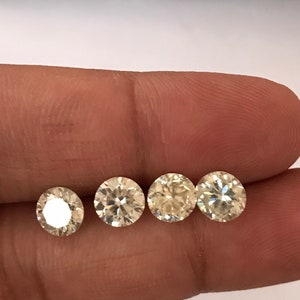 Könnte beinhalten: Vier rund geschliffene Diamanten in verschiedenen Größen werden gezeigt. Die Diamanten werden von einer Hand gehalten, die ihre Brillanz zeigt. Die Steine haben einen warmen, gelblichen Farbton und reflektieren das Licht. Die Diamanten sind vor einem neutralen Hintergrund.