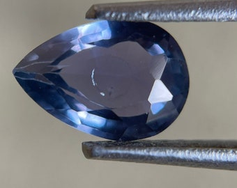 May include: A pear-shaped gemstone with a deep blue color, held in tweezers. The gemstone has a faceted cut and a high degree of brilliance.