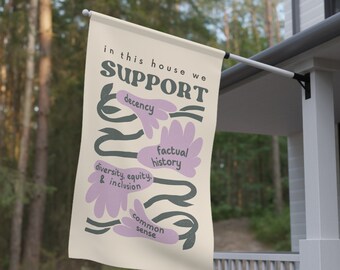 Puede incluir: Una bandera beige con el texto "in this house we SUPPORT" en letras negras. El texto está rodeado de flores estilizadas en morado y verde. Las flores tienen el siguiente texto en negro: "decency", "factual history", "diversity, equity & inclusion", y "common sense".