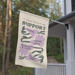 May include: A beige flag with the text "in this house we SUPPORT" in black lettering. The text is surrounded by stylized flowers in purple and green. The flowers have the following text in black: "decency", "factual history", "diversity, equity & inclusion", and "common sense".