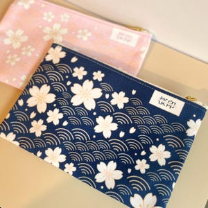 Könnte beinhalten: Zwei Reißverschlusstaschen mit floralen Mustern. Eine ist marineblau mit weißen Kirschblüten und Wellendesigns. Die andere ist rosa mit weißen Kirschblüten. Beide haben goldene Reißverschlüsse und ein kleines weißes Etikett mit japanischem Text.