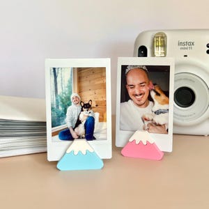 May include: Two white photo frames with pictures of people and dogs, each propped up by a colorful, mountain-shaped stand. One stand is light blue, the other is pink. A white Instax Mini 11 camera is in the background.