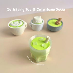 Puede incluir: Una colección de miniaturas de juguetes de comida en verde claro y blanco, que incluyen un matcha latte, una taza pequeña con un sombrero blanco, un cuenco con un batidor y un vaso de matcha helado. El texto "Satisfying Toy & Cute Home Decor" está arriba.