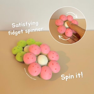 May include: A flower-shaped fidget spinner with pink petals and a white center, accompanied by a second green version. The text reads Satisfying fidget spinner and Spin it! A circular inset shows the pink spinner being rotated by a finger.