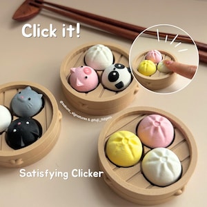 May include: Three round, light brown bamboo-style steamer baskets containing various colourful, bun-shaped fidget toys. The toys include a white bun, a pink pig, a black and white panda, a grey cat, a black cat, a yellow bun, and a pink bun. Text reads Click it! and Satisfying Clicker.