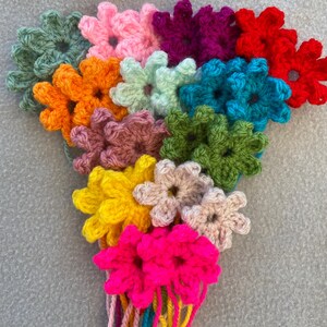 May include: A collection of handmade, crocheted flowers in a variety of colours, including pink, green, orange, yellow, and blue. The flowers are grouped together with yarn strands, creating a decorative floral display.