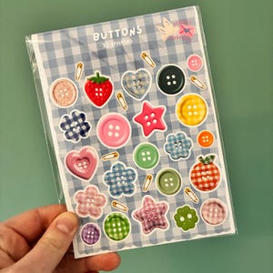 Puede incluir: Una lámina de pegatinas 3D con varios diseños de botones. Las pegatinas incluyen formas como corazones, estrellas y flores, junto con botones redondos clásicos. La lámina está empaquetada en plástico transparente y tiene la palabra "BUTTONS" impresa.