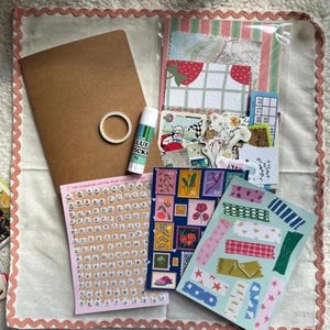 May include: A selection of scrapbooking materials, featuring a brown notebook, glue stick, washi tape, stickers, and patterned paper. The items are arranged on a white surface with a pink scalloped border. The image includes the text "GLUE TICKS".