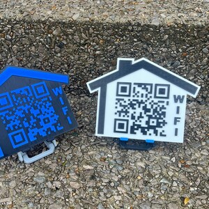 May include: Two house-shaped WiFi signs with QR codes. One is black with a blue outline, the other is white with a black outline. Both have the word "WIFI" printed vertically. They have a small handle at the bottom.