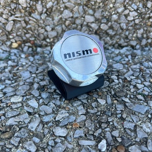 May include: A silver-colored Nismo oil cap with the Nismo logo in black and red. The cap is sitting on a black base. The background is a textured surface of small gray stones.